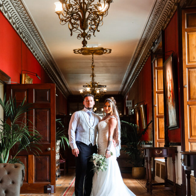 Bride and groom posing under a Victorian chandelier in the corridor of Swinton Park Estate Hotel, luxury Yorkshire wedding.